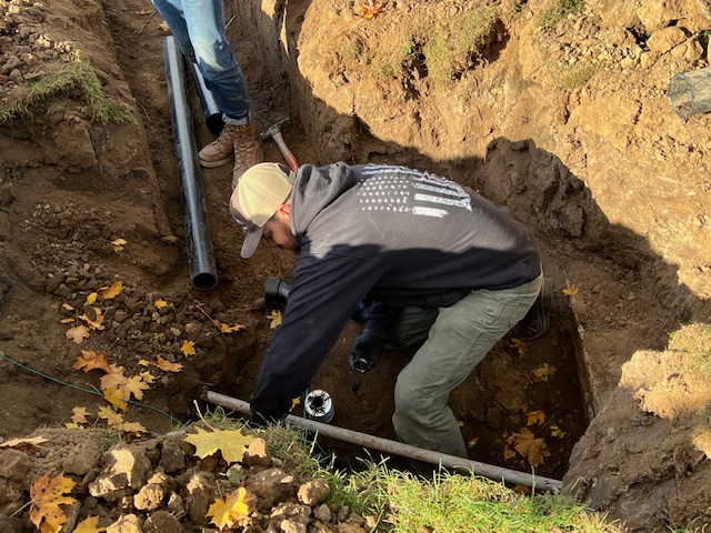 Two people work in a dirt trench with pipes and tools, surrounded by fallen leaves. One person is kneeling and adjusting a pipe fitting.