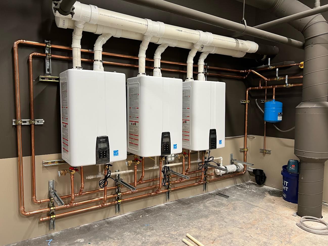 Three wall-mounted tankless water heaters with connected copper piping and white PVC venting, set against a beige and brown wall in a utility room.