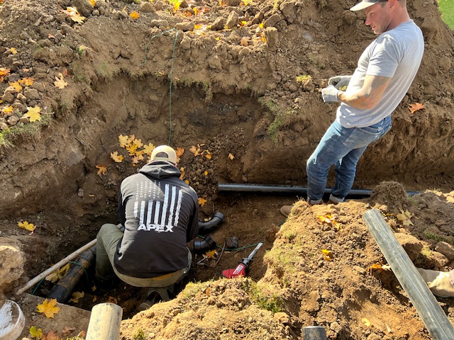 Two workers in a dirt trench install or repair underground pipes; one is kneeling and working, while the other stands nearby holding tools.