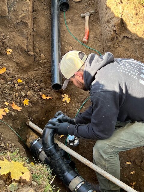 A person wearing gloves installs black plastic plumbing pipes in a dirt trench outdoors, surrounded by fallen autumn leaves and tools.