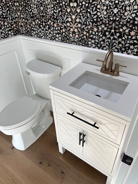 A small bathroom with a white toilet, white vanity with geometric design, brass faucet, wood floor, and floral wallpaper above white wainscoting.