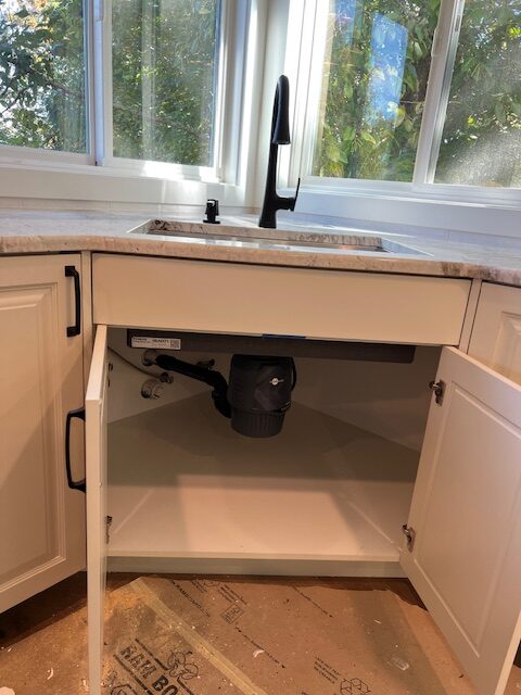 White kitchen cabinets with open doors under a sink, showing exposed plumbing and a garbage disposal unit, with sunlight coming through windows above.