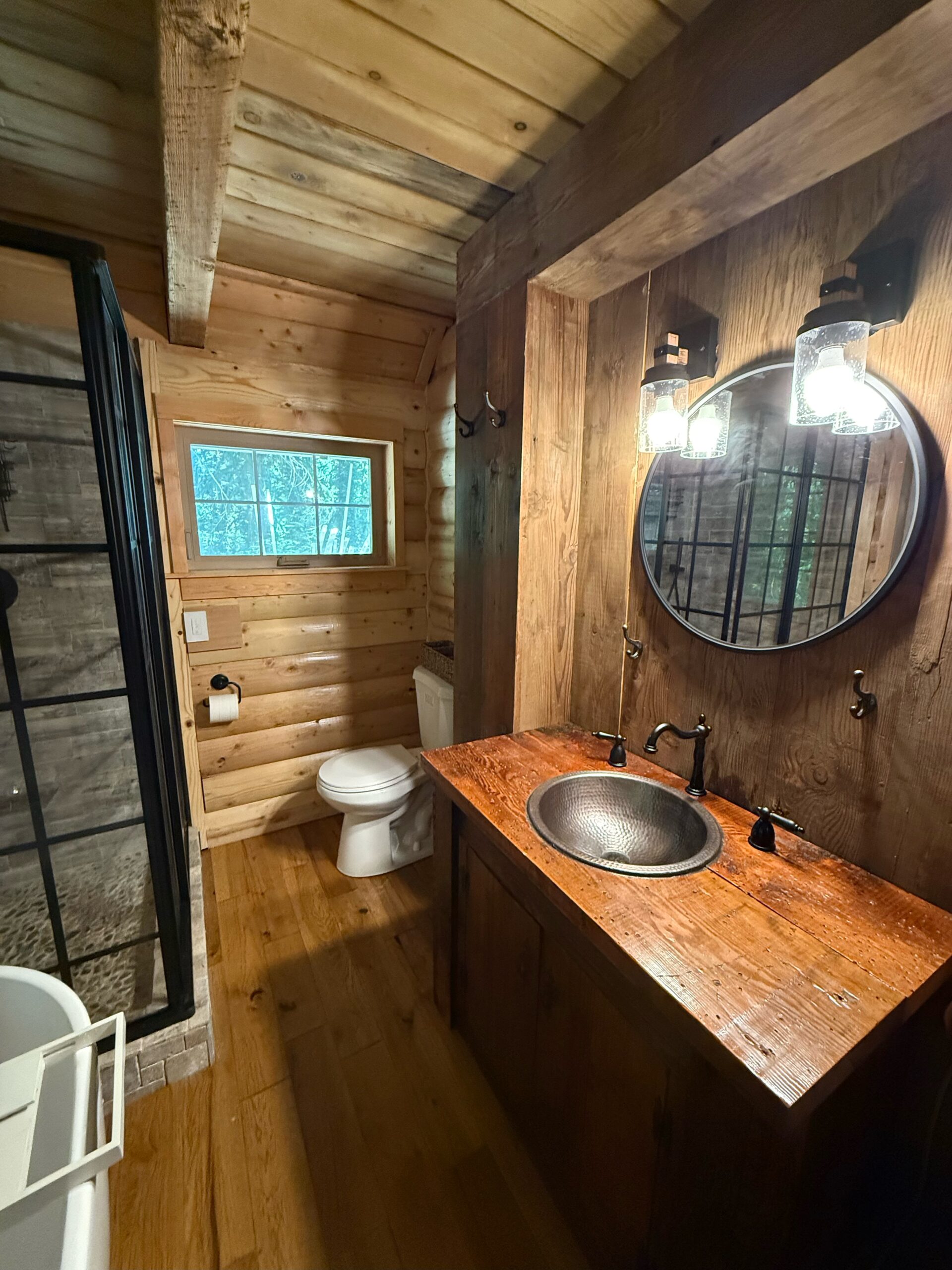 A rustic bathroom with wood-paneled walls, a round mirror above a wooden vanity, black fixtures, a toilet, a window, and a glass shower enclosure.