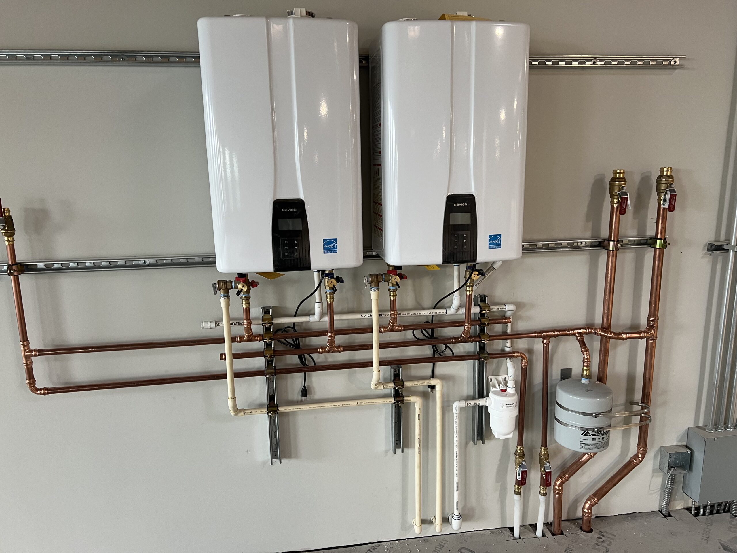 Two wall-mounted tankless water heaters with connected copper and PVC piping, a water filter, and an expansion tank installed on a gray wall.