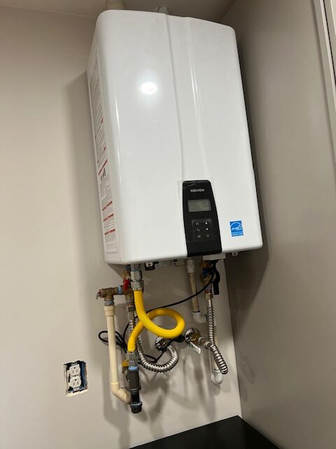 A white tankless water heater is mounted on a wall, with various pipes and a yellow flexible gas line connected below it. An uncovered electrical outlet is nearby.