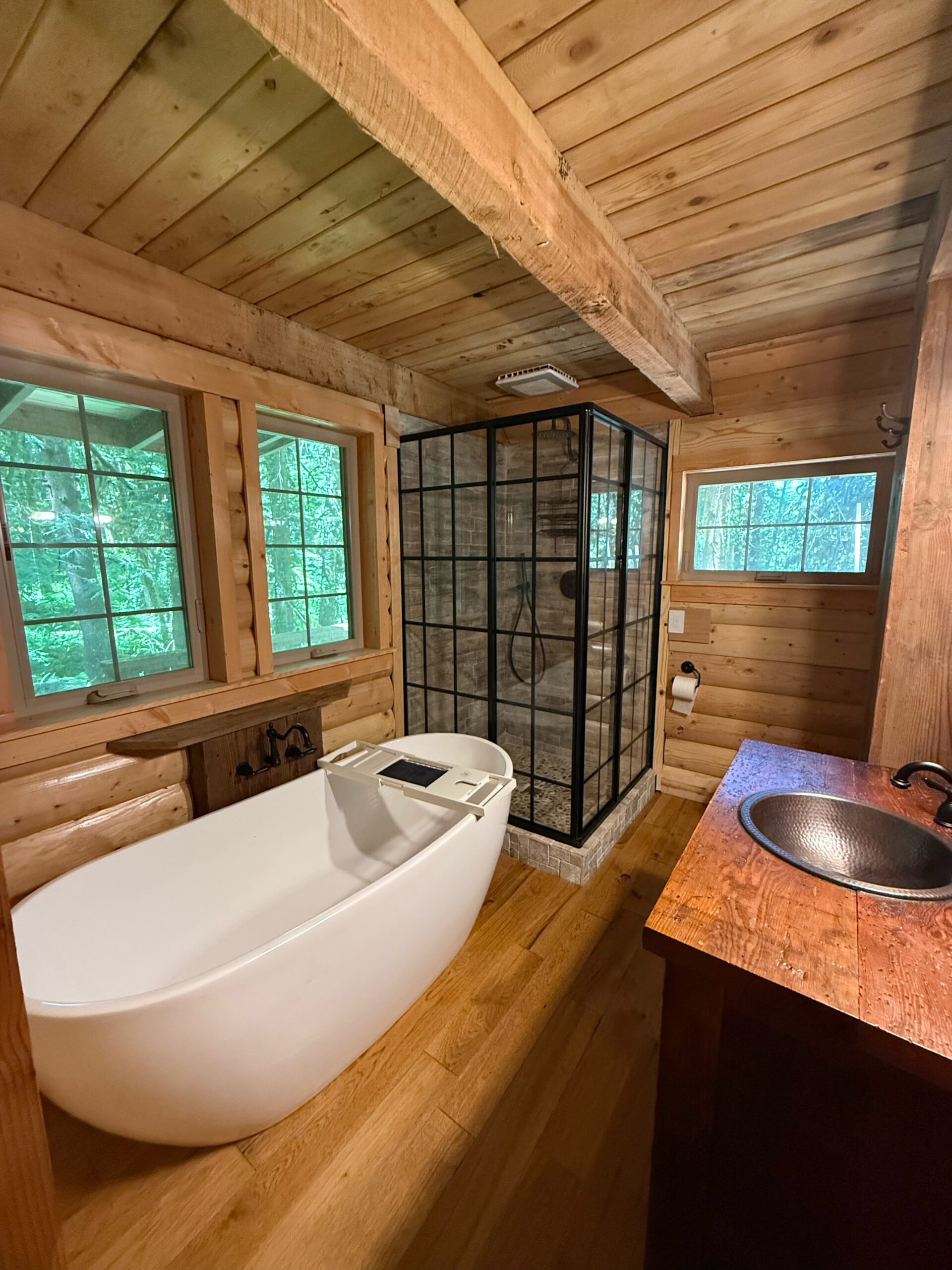 A rustic bathroom with a freestanding white tub, a glass-enclosed shower, wood walls and ceiling, a metal sink, and large windows overlooking trees.